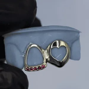 Custom gold two-tooth grillz: one open-face cap with ruby pavé base, one heart-shaped cutout fang cap, shown on a dental mold